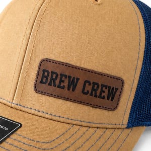 May include: A tan baseball cap with a dark brown leather patch that reads "BREW CREW" in bold, black letters. The cap has a navy blue mesh back and a curved brim. The stitching is a light blue color.