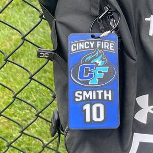 May include: A blue and white Cincy Fire team tag with the name "SMITH" and the number "10". The tag is attached to a black backpack with a silver carabiner. The background includes a baseball bat and a chain-link fence.