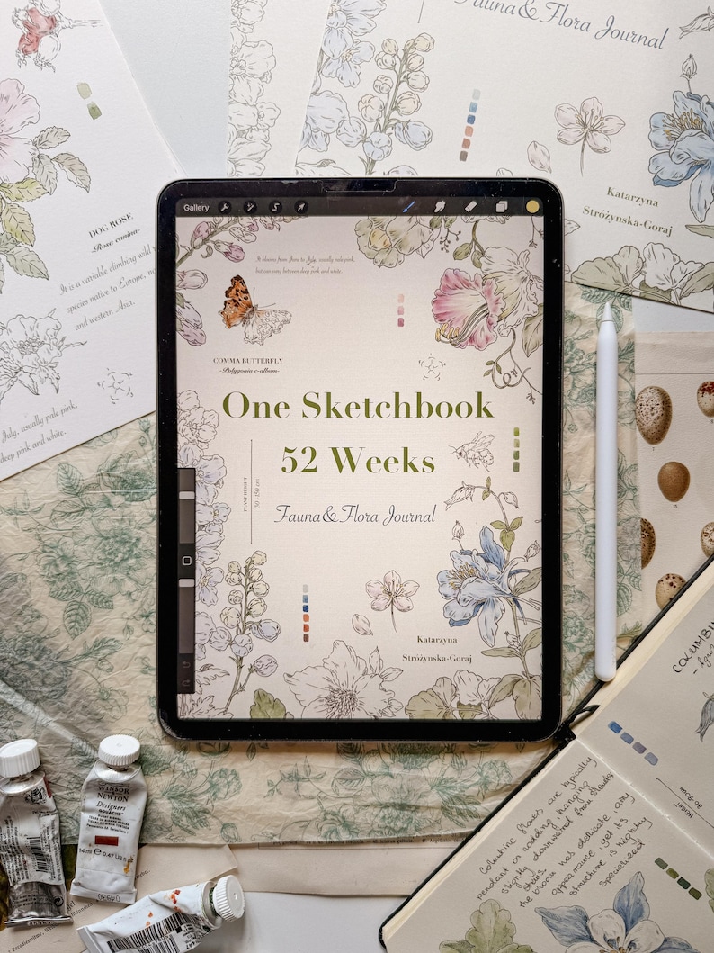 May include: A digital tablet displaying a cover titled One Sketchbook 52 Weeks Fauna & Flora Journal by Katarzyna Str&oacute;żyńska-Goraj, featuring botanical and insect illustrations in soft colors, accompanied by a white stylus and paint tubes.