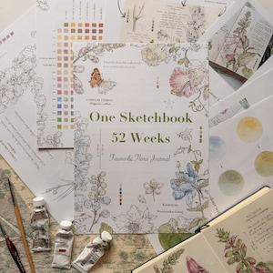 May include: A sketchbook titled One Sketchbook 52 Weeks Fauna & Flora Journal by Katarzyna Strzynska-Goraj, featuring botanical illustrations of flowers and a butterfly, surrounded by watercolor paint tubes, brushes, and instructional pages.