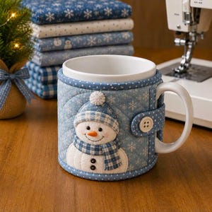May include: A white ceramic mug featuring a removable quilted fabric cozy in light blue with a white snowflake pattern. The cozy displays a plush snowman applique wearing a blue plaid hat and scarf, secured by a single white button.