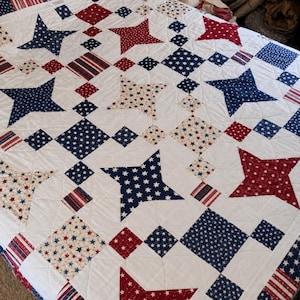 May include: A white quilt with a patriotic design featuring red, white, and blue stars and diamond shapes. The quilt has a patchwork pattern with various star sizes and striped accents, creating a classic Americana theme.