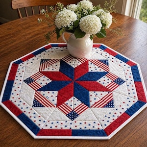 May include: An octagonal patriotic table topper with a starburst design. The red, white, and blue quilt features American flag and star patterns. A white pitcher with white flowers sits on the topper, which is on a wooden table.