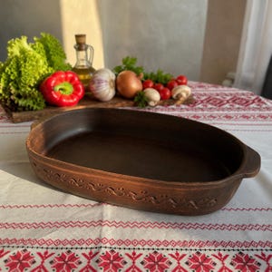 Puede incluir: Una fuente de horno ovalada de arcilla marrón con asas laterales integradas y un patrón decorativo de ondas en relieve a lo largo del borde exterior. El interior es liso y oscuro, mientras que el exterior tiene una textura natural.