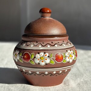 Puede incluir: Un tarro de cerámica marrón con tapa, adornado con un motivo floral. El tarro presenta flores blancas con detalles rojos y verdes, y una banda decorativa. La tapa tiene un pequeño pomo redondeado.