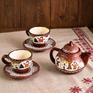 以下が含まれることがあります： 茶色の陶器製ティーセット。カップ2客とソーサー、ティーポットで構成されています。各アイテムには、白、オレンジ、緑で手描きされた冬の風景と、白い水玉模様が施されています。