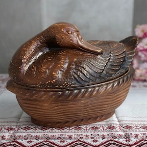 Puede incluir: Un recipiente de cerámica marrón con tapa en forma de pato que descansa sobre una base de cesta tejida. El pato presenta plumas talladas detalladas y una cabeza lisa, mientras que la base tiene un borde texturizado y líneas horizontales.