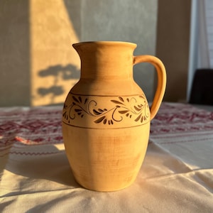 Pode incluir: Uma jarra de terracota em tom castanho claro com boca larga, pega curva e corpo arredondado. A parte superior da jarra apresenta uma faixa horizontal decorada com um padrão esculpido de videira floral repetitiva num tom castanho escuro.