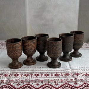May include: A set of six dark brown clay goblets with a textured, wood-grain appearance. Each goblet features a rounded bowl, a short stem, and a circular base. The vessels are arranged in a staggered formation, showcasing their uniform shape.