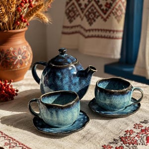 Op de afbeelding: Een keramische theeset bestaande uit een theepot en twee kopjes met bijpassende schoteltjes. De stukken hebben een gespikkeld blauw en zwart glazuur met een glanzende afwerking en onregelmatige, organische vormen.