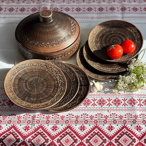 May include: Handmade ceramic tableware set featuring brown plates and a lidded serving dish. The pottery has detailed carved patterns and is displayed on a white tablecloth with a red and white geometric design. Two tomatoes and garlic are also visible.