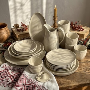 May include: A collection of off-white ceramic dinnerware including a pitcher, various plates, bowls, and cups with irregular, organic edges. The set is arranged on a wooden surface alongside a small candle holder and a patterned textile.