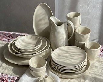 May include: A collection of off-white ceramic dinnerware featuring a pitcher, various plates, bowls, and cups with organic, wavy edges. The set includes a small candle holder with a handle, all finished in a smooth, glossy, light-colored glaze.