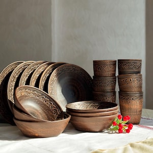 May include: A set of dark brown clay dinnerware including several plates, bowls, and cups. Each piece features a textured surface with a carved, repeating leaf-like pattern along the rim. The items are arranged in a stack and a group.