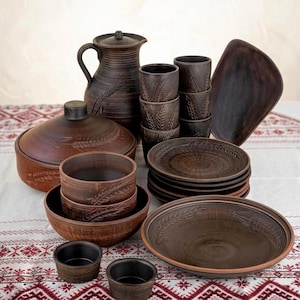 Pode incluir: Um conjunto de louça de barro castanho escuro composto por um jarro com tampa, uma caçarola com tampa, várias taças, chávenas e pratos. Cada peça é decorada com um padrão subtil de espigas de trigo em relevo na superfície exterior.