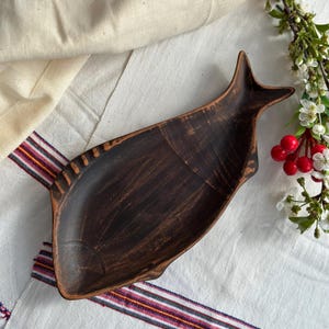 May include: A dark brown, fish-shaped wooden serving tray. The tray has a carved design with a tail and fins. The tray is placed on a white cloth with a striped border. Decorative flowers and berries are in the background.