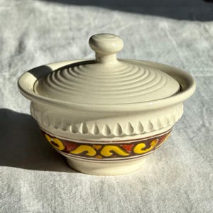 May include: A cream-coloured ceramic sugar bowl with a matching lid featuring a concentric ring pattern. The bowl has a carved teardrop texture around the upper rim and a decorative band of yellow and orange scrollwork on a dark brown background.