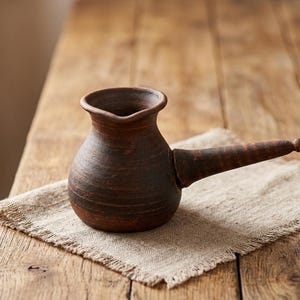 Pode incluir: Uma cafeteira de argila castanho-escura com corpo arredondado, borda larga e alargada, e uma pega longa e cónica que se estende lateralmente. A superfície do recipiente apresenta um acabamento mate texturado com estrias horizontais visíveis.