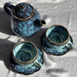 May include: A ceramic tea set with a blue glaze. The set includes a teapot with a lid and two teacups with saucers. The rims of the cups and teapot are a darker shade of blue. The set is on a white surface.