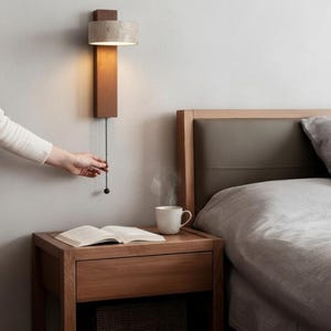 Könnte beinhalten: Eine moderne Wand-Nachttischlampe mit rechteckigem Holzsockel und zylindrischem Betonschirm. Die Lampe wird durch Ziehen an einer Kette eingeschaltet. Ein Nachttisch mit Buch und Kaffeetasse.