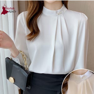 May include: A white long-sleeved blouse featuring a high mock neck collar adorned with three small white faux pearl accents. The front of the blouse displays vertical pleats extending from the neckline, and the cuffs are finished with pearl buttons.