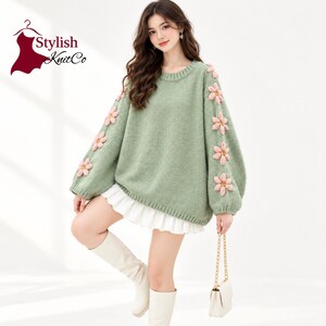 May include: A sage green long-sleeve sweater featuring a crew neckline and ribbed cuffs. The sleeves are decorated with a vertical row of pink, flower-shaped appliques. The Stylish KnitCo logo is visible in the upper left corner of the image.