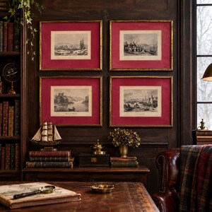 May include: Four rectangular wall art prints in gold frames with deep red matting, each featuring a black and white landscape illustration. The prints are arranged in a grid pattern on a dark wood paneled wall above a wooden desk with books.