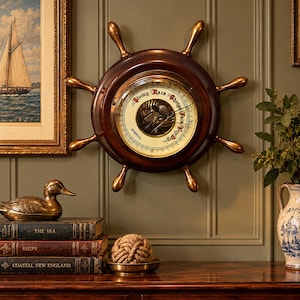 May include: A wall-mounted barometer shaped like a ship's wheel, featuring a dark wood frame, brass handles, and a cream-colored dial with the text Stormy, Rain, Change, Fair, Very Dry, and Gabros printed in black lettering.