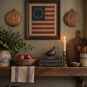Könnte beinhalten: Eine gerahmte amerikanische Flagge mit einem Sternenkreis, zwei kupferne Adler-Wandplatten, eine Holzschale mit roten Äpfeln, ein Stapel Bücher mit den Titeln American Antiques, Colonial Home und New England, ein Messingkerzenhalter und ein gestreiftes Tuch.