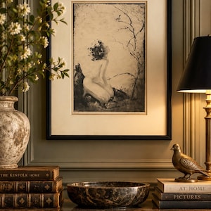 Peut inclure: Une impression en noir et blanc encadrée représentant une silhouette agenouillée sous un arbre dénudé, présentée dans un cadre noir avec un passe-partout crème. L'œuvre est placée au-dessus de livres, d'un bol en marbre et d'une figurine d'oiseau.