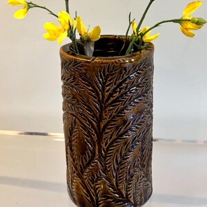 May include: A cylindrical brown ceramic vase featuring a textured, embossed fern leaf pattern across its exterior surface. The vase contains several thin green stems with small, bright yellow flowers extending from the top opening.