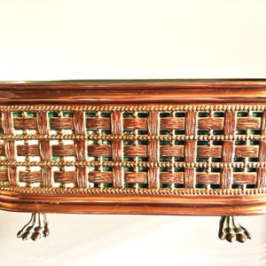 May include: A rectangular copper planter with a woven basket-weave pattern and a green interior lining. The vessel features a beaded border along the top and bottom edges and is supported by four metallic claw-style feet at the base.