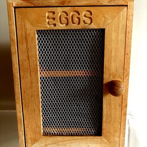 May include: A rectangular light brown wooden egg storage cabinet featuring a single hinged door with a round wooden knob. The door contains a silver-coloured metal wire mesh panel and the word EGGS is carved into the top wooden frame section.
