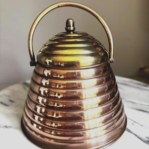 Könnte beinhalten: Ein bienenkorbförmiger Behälter aus poliertem Kupfer mit einem Messingdeckel und einem passenden schwenkbaren Messinggriff. Der Körper weist durchgehend horizontale Rippendetails auf, die ein gestuftes Erscheinungsbild bis zum Knauf bilden.