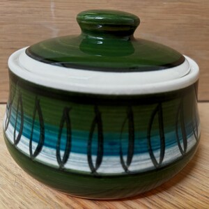 Può includere: Una zuccheriera rotonda in ceramica con coperchio abbinato, caratterizzata da fasce orizzontali color verde bosco, verde acqua e bianco. L'esterno è decorato con un motivo ripetitivo di contorni neri a forma di foglia dipinti a mano.