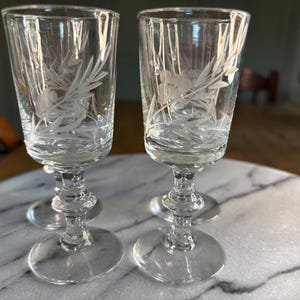 Op de afbeelding: Twee heldere glazen op voet met een geëtst bloemmotief van bladeren en bessen op de kelk. Elk glas heeft een decoratieve steel met meerdere niveaus en een ronde voet, wat zorgt voor een stabiele basis voor het hoge, slanke drinkglas.