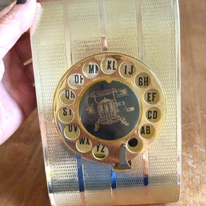 Könnte beinhalten: Ein goldfarbenes, rechteckiges Telefonregister mit strukturierter Oberfläche und einem kreisförmigen Drehscheibenmechanismus. In der Mitte der Wählscheibe befindet sich eine schwarze grafische Abbildung einer alten Maschine, umgeben von Buchstabenpaaren von AB bis YZ.