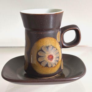 May include: A dark brown ceramic cup and saucer set featuring a white interior and a prominent mustard yellow circular pattern with a stylized flower motif in the center. The cup has a cylindrical body with a thick handle and a square-shaped saucer.
