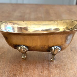 Pode incluir: Uma pequena taça de latão polido em forma de banheira em miniatura com pés. O recipiente apresenta um rebordo curvo e liso, apoiando-se em quatro pés distintos, cada um concebido com um motivo decorativo em forma de concha no topo.