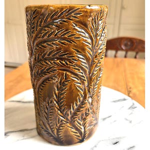 May include: A cylindrical ceramic vase with a glossy brown glaze and a deep relief pattern of fern-like leaves. The surface features dark brown accents within the recessed areas of the botanical design, creating a textured appearance on the exterior.