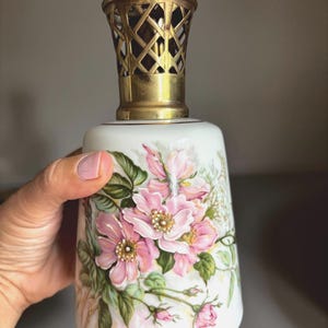 May include: A white ceramic fragrance lamp featuring a hand-painted floral design with pink blossoms, green leaves, and gold accents. The top of the vessel is fitted with a decorative, perforated gold-tone metal crown cap with a lattice pattern.