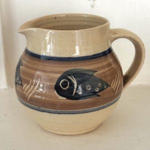 May include: A beige ceramic pitcher with a rounded body and a single handle. The exterior features a wide horizontal band in a light brown tone, decorated with dark blue fish motifs and white wavy lines, accented by thin blue horizontal stripes.
