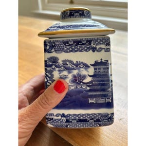 May include: A square ceramic canister with a lid featuring a blue and white willow pattern. The design depicts a traditional pagoda, trees, and water landscape. The lid and the top rim of the canister are accented with a thin gold metallic trim.