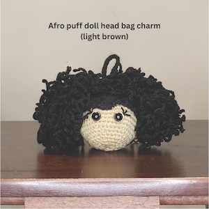May include: A crochet bag charm featuring a light brown doll head with black afro puff hair. The text above the charm reads: Afro puff doll head bag charm (light brown). The doll has black plastic eyes and stitched eyelashes.