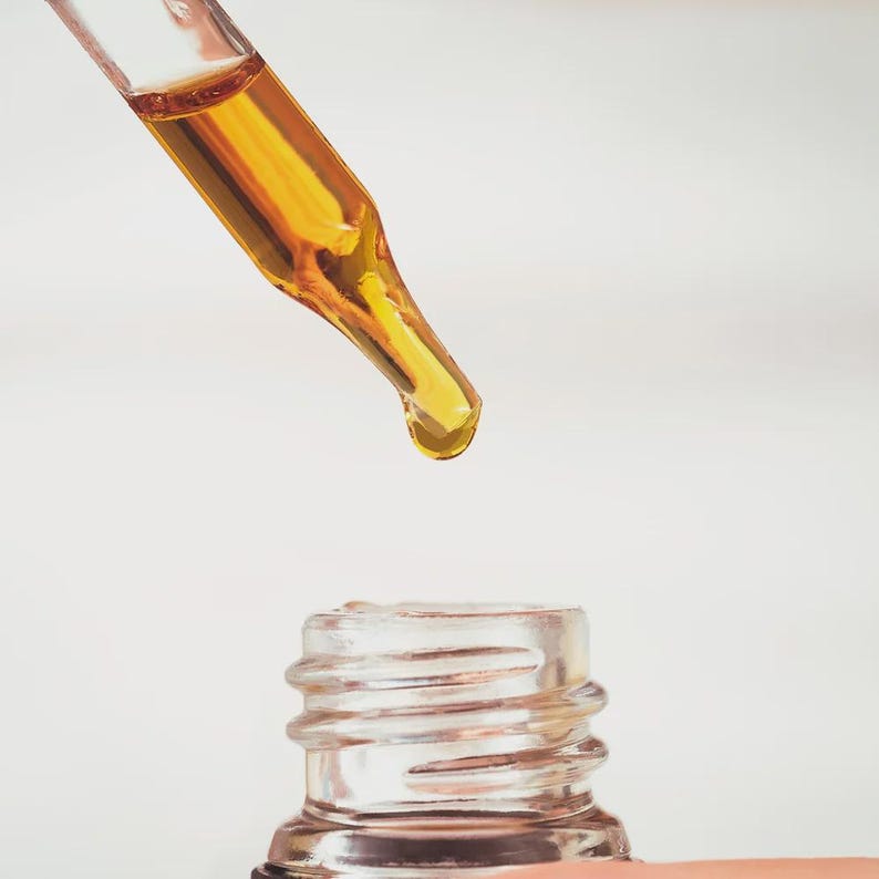 May include: Close-up of a glass dropper dispensing a golden liquid into a small, clear glass bottle. The bottle has a ribbed neck. The background is a soft, neutral white.