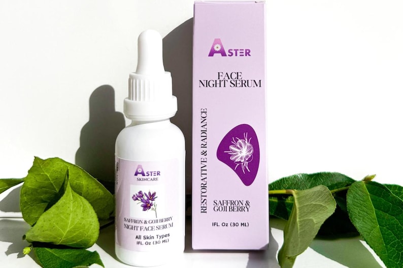 May include: A white bottle of Aster Skincare Face Night Serum with a dropper, next to a matching box. The box and label feature the text "Saffron & Goji Berry" and a purple floral design. Green leaves are in the foreground.