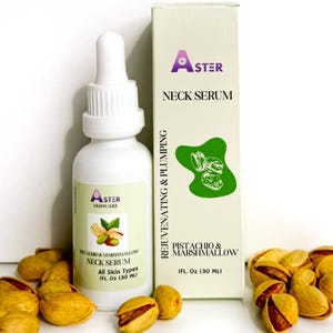May include: A white bottle of Aster Skincare Neck Serum with a dropper, next to a box with the same branding. The box and bottle are labeled "Pistachio & Marshmallow." Shelled pistachios are scattered around the bottle and box.