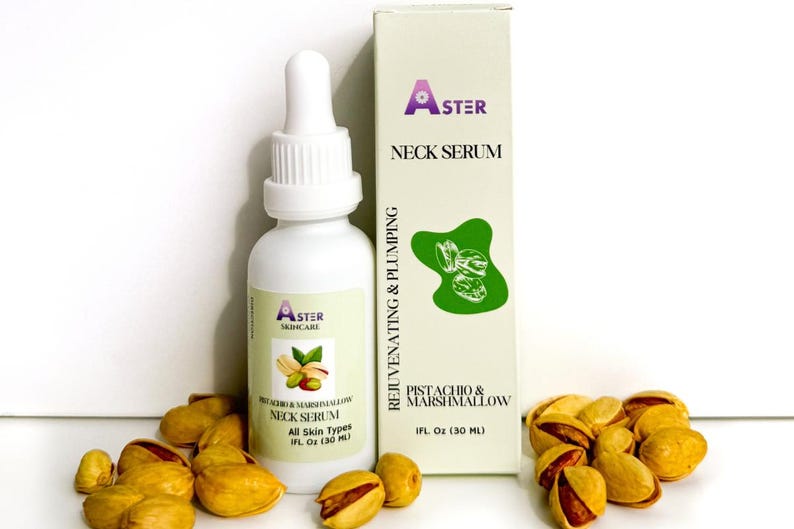 May include: A white bottle of Aster Skincare Neck Serum with a dropper, next to a box with the same branding. The box and bottle are labeled "Pistachio & Marshmallow." Shelled pistachios are scattered around the bottle and box.