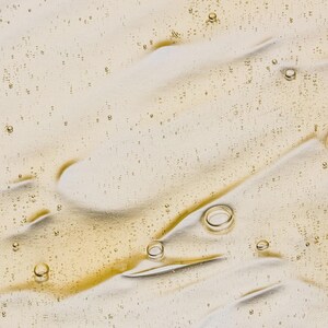 May include: Close-up of a light beige liquid with a glossy texture. The surface is marked with streaks and small, scattered bubbles. The liquid has a slightly golden hue, suggesting a cosmetic or skincare product.