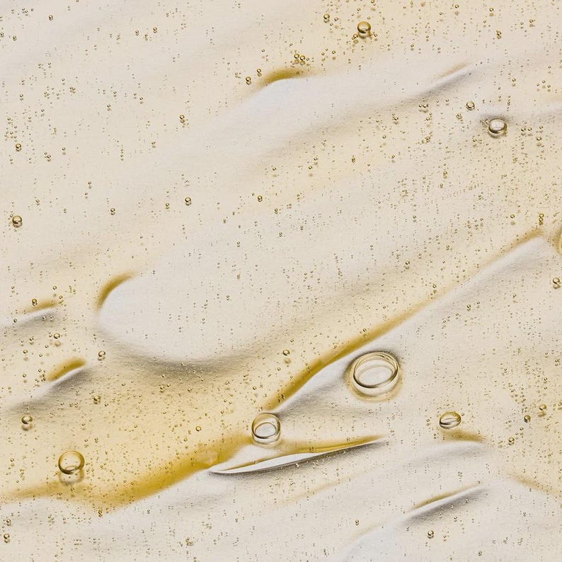 May include: Close-up of a light beige liquid with a glossy texture. The surface is marked with streaks and small, scattered bubbles. The liquid has a slightly golden hue, suggesting a cosmetic or skincare product.